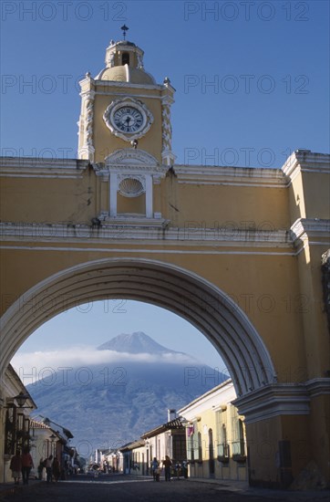GUATEMALA, Antigua, "Arco de Santa Catarina. Yellow painted archway with clock tower and people walking, with Volcan Agua visible through arch."