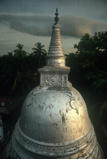 SRI LANKA, Anuradhapura, "Carved Dagoba near Isurumuniya rock temple, damaged surface shining silver / white in low light, surrounded by trees."