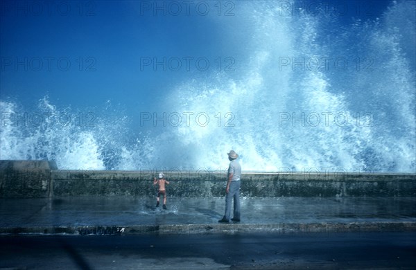 CUBA, Havana, Malecon, Old man and small boy standing on pavement with waves crashing over the sea wall