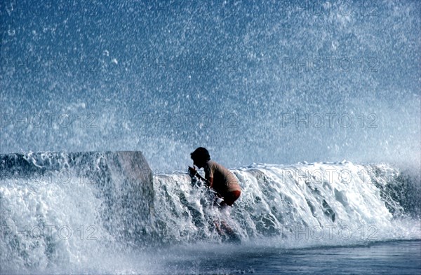 CUBA, Havana, Malecon, Young boy standing on pavement with waves crashing over the sea wall