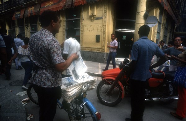 CUBA, Havana, Newspaper vendor selling goods from a bicycle seat in the street with passing traffic and people