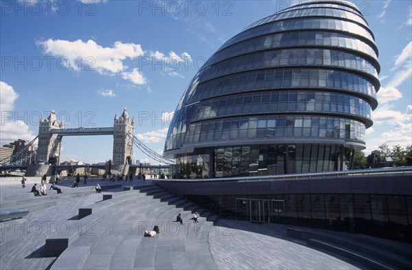 ENGLAND, London, City Hall exterior view of the curved architecture with Tower Bridge in the distance and people sitting on the steps in the foreground