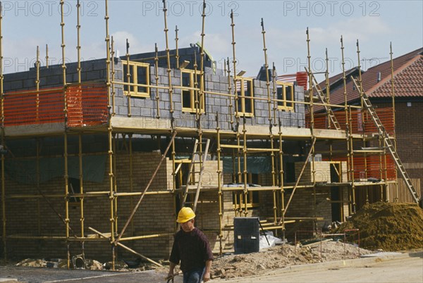 ARCHITECTURE, Construction, Building Site, Scaffolding surrounding half completed brick house with man wearing a yellow hard hat in the foreground