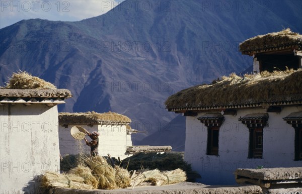 CHINA, Tibet, Yanjing, Farmer winnowing barley by mountain village farm buildings