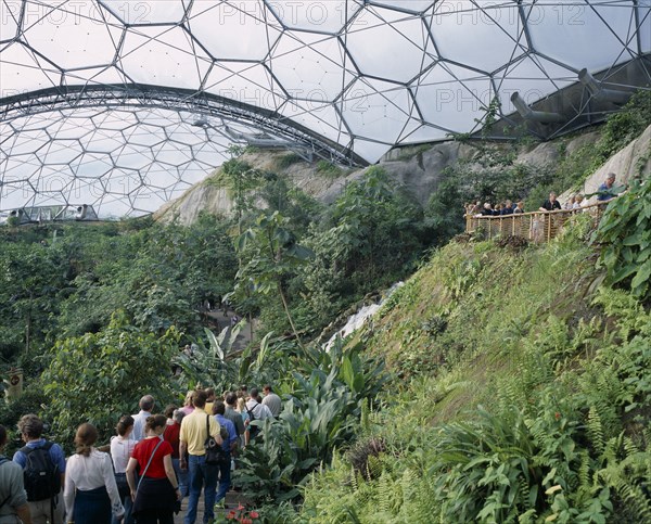 ENGLAND, Cornwall, St Austell, Eden Project. Tropical dome interior with visitors on path leading past waterfall