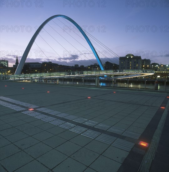 ENGLAND, Tyne and wear, Gateshead, The new Millennium Footbridge illuminated at dusk with city buildings silhouetted against purple evening sky behind.