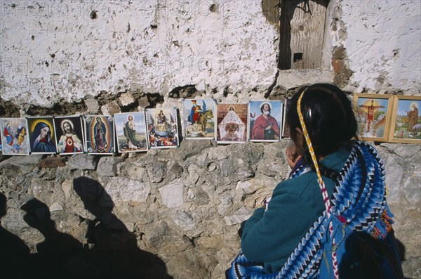 MEXICO, Chiapas, San Cristobal de Las Casas, Woman in front of coloured pictures of the lives of the Saints and Christ displayed along stone wall.