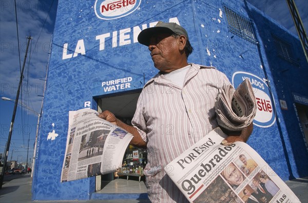 MEXICO, Yucatan, Merida, "Male newspaper vendor, threequarter view seen from below looking up.  Advertising for Nestle and purified water on blue painted wall of building behind. "