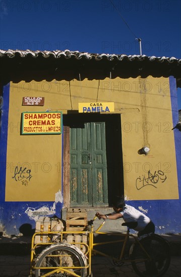 MEXICO, Chiapas, San Cristobal de Las Casas, Man pushing bicycle handcart stacked with crates of fruit past yellow and blue painted building facade with advertisment for shampoo beside green door.