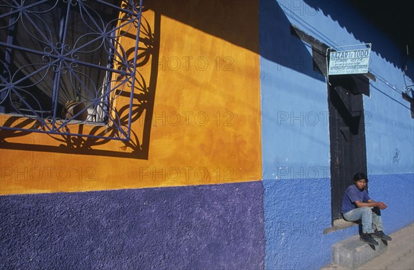 MEXICO, Chiapas, San Cristobal de Las Casas, "Part view of building painted in rectangles of ochre, blue and purple with partly open purple metal window screen  casting shadow.  Boy sitting in doorway."