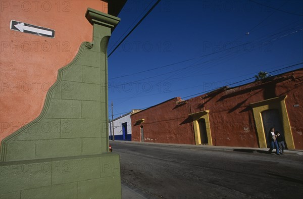 MEXICO, Oaxaca, Oxaca City, Corner of green and orange painted building with white arrow on black background pointing left.  Long orange building with yellow door frames across road behind.