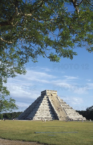 MEXICO, Yucatan, Chichen Itza, "El Castillo, angled view of facade partly framed by tree branches with visitors climbing steep steps to the top."