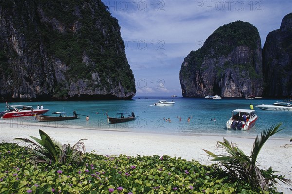 THAILAND, Krabi Province, Ko Phi Phi Leh , Maya Bay.  View over purple flowers towards white sand beach overlooked by limestone rock formations.  Line of long tail and tourist boats and people swimming.