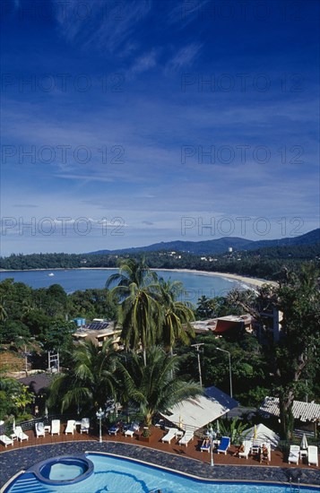 THAILAND, Phuket , Kata Beach, View over hotel swimming pool with tourists on sun loungers beside it towards curving sandy beach and distant tree covered hillside.