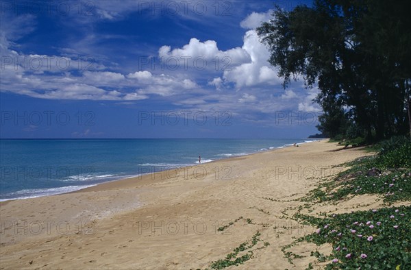 THAILAND, Phuket , Mai Khao Beach, View along quiet sandy beach fringed with trees and vegetation with people in the water in the distance.