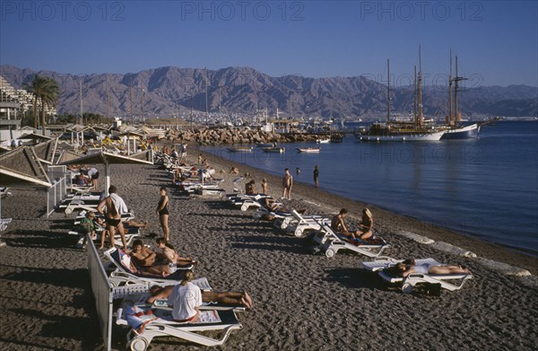 ISRAEL, Eilat, Coral Beach, "Lines of sunbathers on white loungers on dark, sand beach with sailing ships moored against jetty behind and distant mountain range."