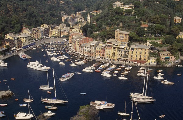 ITALY, Liguria, Portofino, Aerial view over harbour with moored yachts and pastel coloured town houses with tree covered hillside behind.