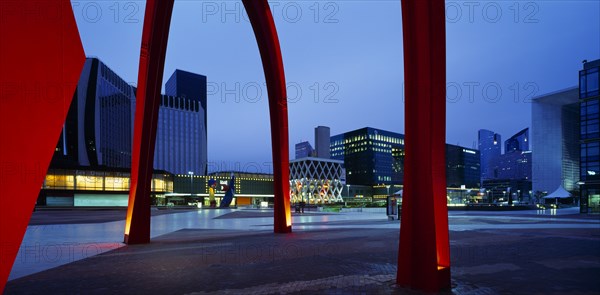 FRANCE, Ile de France, Paris, La Defense.  Alexander Calders red iron stabile sculpture.  Part view with city buildings behind illuminated at night.