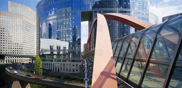 FRANCE, Ile de France, Paris, "La Defense.  Covered pedestrian bridge over busy road with circular, glass fronted building reflecting city skyline behind."