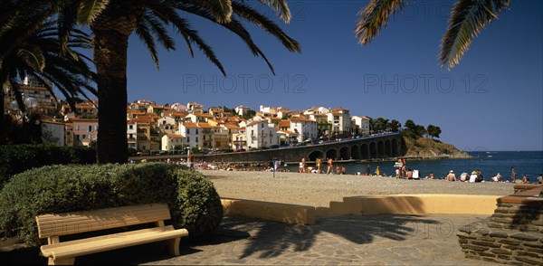 FRANCE, Languedoc-Roussillon, Pyrenees Orientals, Banyuls-Sur-Mere.  View across paved area and pebble beach with sunbathers towards town houses on hill behind.