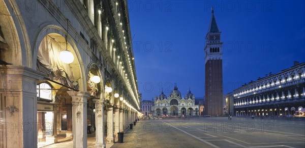 ITALY, Veneto, Venice, Piazza San Marco.  View along the colonnaded Procuratie Vecchie towards the Basilica di San Marco and the Campanile illuminated at dusk.