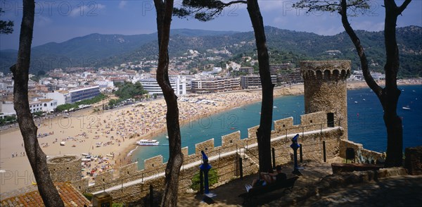 SPAIN, Catalonia, Tossa de Mar, View over busy sandy beach and town from fortified walls of Villa Vella with line of trees in the foreground.