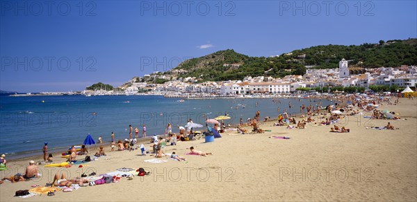 SPAIN, Catalonia, El Port de La Selva, "Curved, sandy beach with people sunbathing and in the water with white painted town at foot of tree covered hillside beyond. "