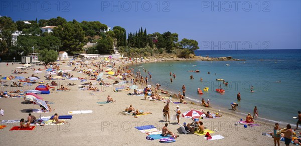 SPAIN, Catalonia, Llanca, "Curved, sandy beach with people sunbathing and in the sea.  White painted houses amongst trees on hillside beyond."