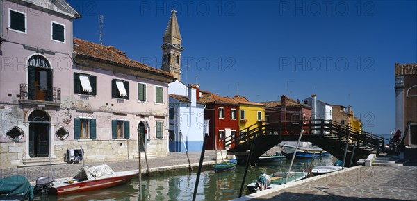 ITALY, Veneto, Venice, "Burano Island.  Bridge and canal with boats moored to posts along each side overlooked by pink, blue, red and yellow buildings and bell tower behind."