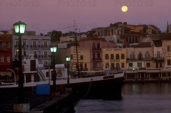 GREECE, Crete, Hania, Venetian Port.  Moored boat and quayside buildings with moon in pale purple dusk sky above and illuminated street lamps.