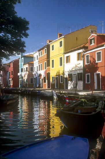 ITALY, Veneto, Venice, Burano Island.  Boats moored at canal-side overlooked by row  of brightly painted buildings.