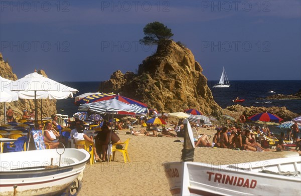 SPAIN, Catalonia, Platja de la Mar Menuda, Busy sandy beach with people sitting at tables under umbrellas or sunbathing.  Central rocky outcrop and boats on beach in foreground.