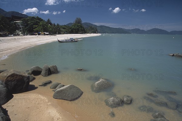 MALAYSIA, Penang, Batu Ferringhi, View along shore of quiet sandy beach with moored boat in middle distance and smooth rocks in shallow water in the foreground.