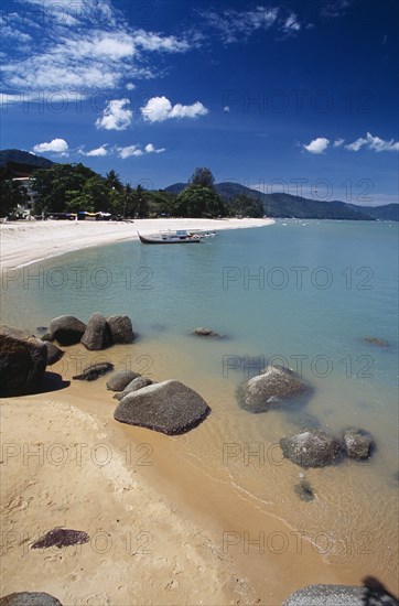 MALAYSIA, Penang, Batu Ferringhi, View along shore of quiet sandy beach with moored boat in middle distance and smooth rocks in shallow water in the foreground.