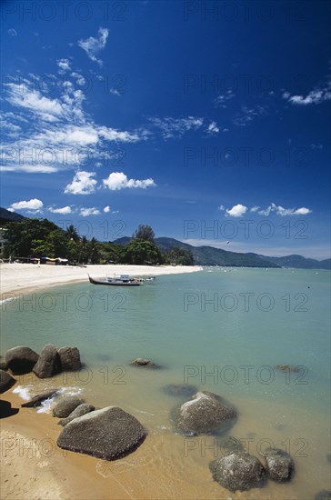 MALAYSIA, Penang, Batu Ferringhi, View along shore of quiet sandy beach with moored boat in middle distance and smooth rocks in shallow water in the foreground.