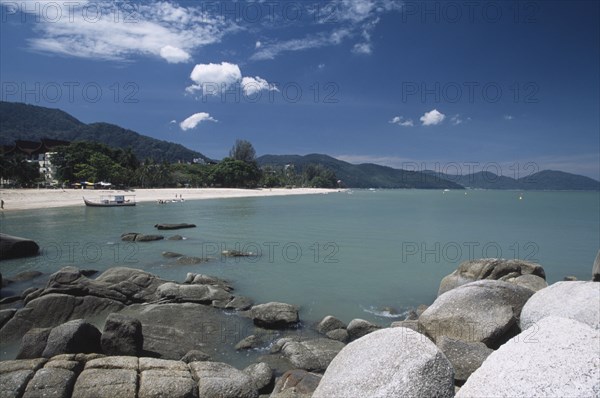 MALAYSIA, Penang, Batu Ferringhi, View over rocks and bay of turquoise water towards distant moored boat and quiet sandy beach.