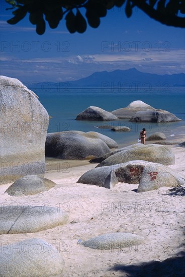 MALAYSIA, Penang, Batu Ferringhi, "Sandy beach with woman in bikini walking out of sea between large, smooth rocks, partly framed by silhouetted tree branch"