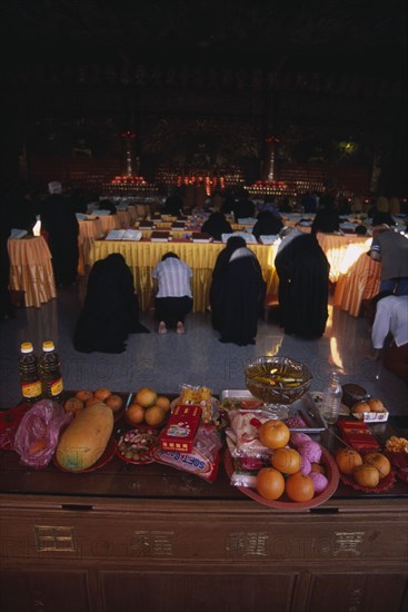 MALAYSIA, Penang, Kek Lok Si Temple, Morning prayers in the main prayer hall. Kneeling worshippers with table in foreground spread with offerings of fruit flowers and candles.