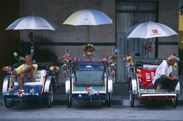 MALAYSIA, Penang, Georgetown, General.  Street scene with three bicycle rickshaws decorated with flowers and flags parked beside roadside kerb with resting drivers.