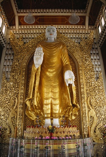 MALAYSIA, Penang, Georgetown, Dharmikarama Burmese Temple.  Interior with large standing Buddha figure with raised hand in gold painted robe and ornate carved gold panel behind.