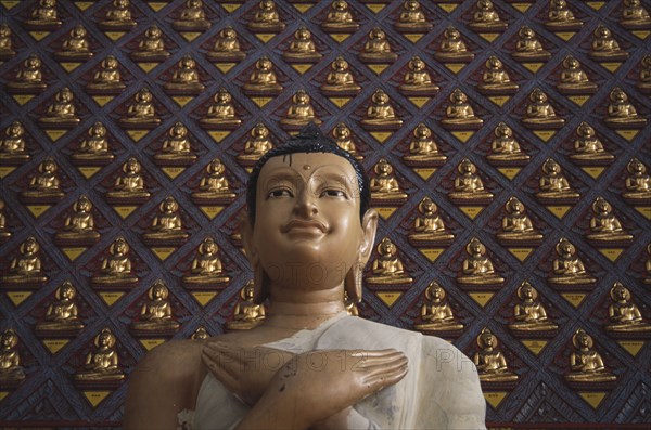 MALAYSIA, Penang, Georgetown, Wat Chayamangkalaram.  Detail of Buddha figure with hands crossed over chest.  Wall behind covered with small raised seated Buddha figures painted gold.