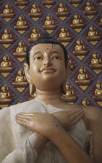 MALAYSIA, Penang, Georgetown, Wat Chayamangkalaram.  Detail of Buddha figure with hands crossed over chest.  Wall behind covered with small raised seated Buddha figures painted gold.