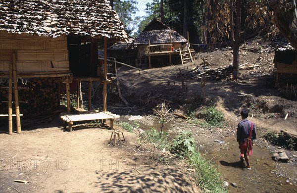 THAILAND, North, Mae Sariang, Young Karen refugee man walking in shallow stream toward wood and cane houses built on stilts