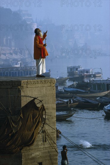INDIA, Uttar Pradesh, Varanasi, "Holy man praying on stone wall above River Ganges,boats,town,mist "