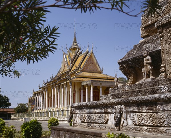 CAMBODIA, Phnom Penh, Silver Pagoda.  Exterior with ornate golden roof with spire and stone monument in foreground.