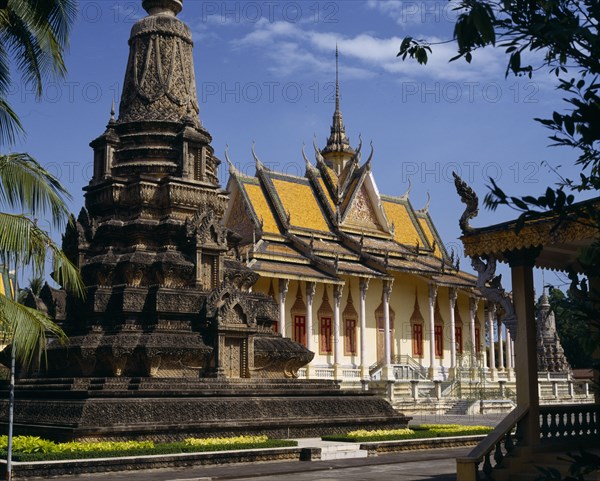 CAMBODIA, Phnom Penh, Silver Pagoda.  Exterior with ornate golden roof with spire and stone monument in foreground.