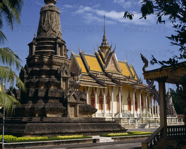 CAMBODIA, Phnom Penh, Silver Pagoda. Exterior with ornate golden roof with spire and stone monument in foreground.