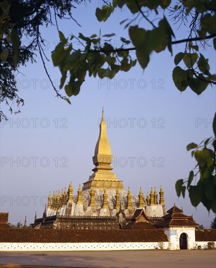 LAOS, Vientiane, Pha That Luang Sacred Stupa with gold spires and white walls part framed by tree branches.