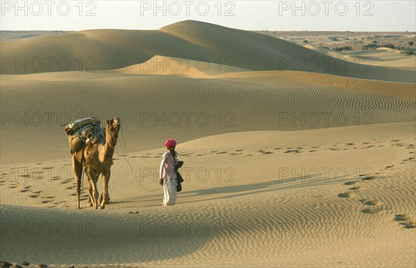 INDIA, Rajasthan, Jaisalmer, Thar Desert.  Man walking with camel carrying pack over dunes leaving prints in the sand.