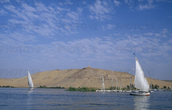 EGYPT, Upper Egypt, Aswan, Qubbet el-Hawa and The Tombs of The Nobles with two feluccas sailing past on the river Nile